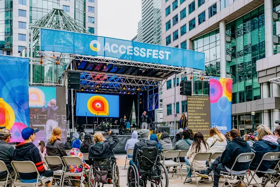 Une foule diversifiée, comprenant des personnes en fauteuil roulant, assiste à un spectacle en direct sur une scène extérieure du centre-ville de Toronto, entourée d'immeubles de bureaux.