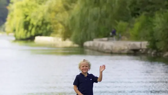 A young child waves from a wooden dock at Bayfront Park in Hamilton, Ontario, with willow trees and calm water in the background.