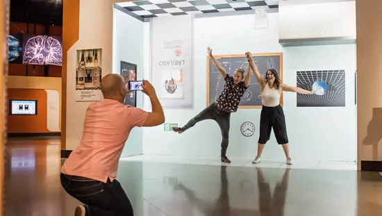 Two visitors strike playful poses in a Lab exhibit at the Canada Science and Technology Museum while a third person photographs them