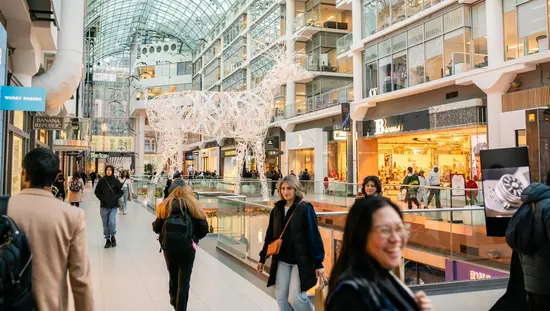 Shoppers fill Toronto's Eaton Centre mall during the holiday season, with large illuminated reindeer sculptures under the iconic vaulted glass ceiling.