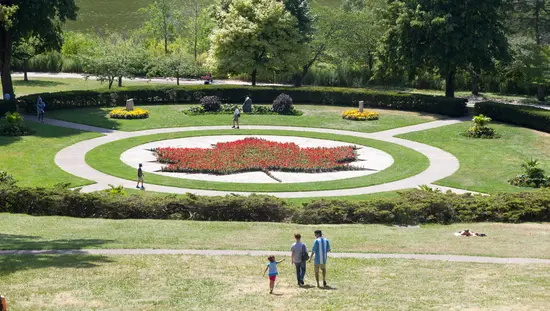 Aerial view of High Park's formal garden featuring a red maple leaf floral display, with a family walking toward it and a pond visible through the trees