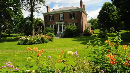 Casa histórica de ladrillo con porche ribeteado en blanco, rodeada de jardines y coloridas flores sobre un césped verde.