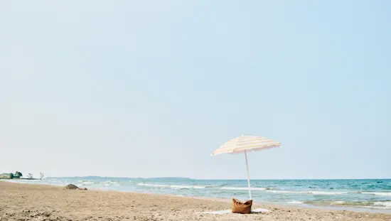 Sandy beach with a striped umbrella and tote bag by the water's edge.