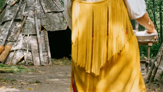 A woman carries a plate to a traditional Indigenous dwelling.