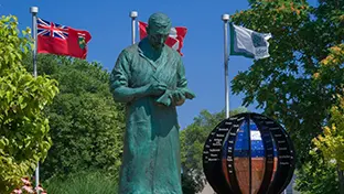 Bronzestatue von Sir Frederick Grant Banting auf einem Steinsockel, dahinter Flaggen und eine Globusskulptur in einem Garten.