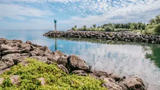 Partially cloudy sky is reflected on the body of water with large rocks where the water meets the grass. 