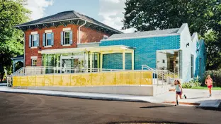 Zwei Kinder rennen auf dem Bürgersteig weg von den braunen und blauen Gebäuden des Hamilton Kindermuseums.