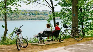 Deux personnes ayant garé leur vélo le long d’un sentier et assises sur un banc noir, d’où elles contemplent un lac avec des arbres en arrière-plan.