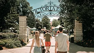 Menschen gehen unter einem kunstvoll verzierten Eisenbogen mit der Aufschrift „Victoria Park“ hindurch in Richtung eines Festivals mit Zelten und Verkaufsständen.