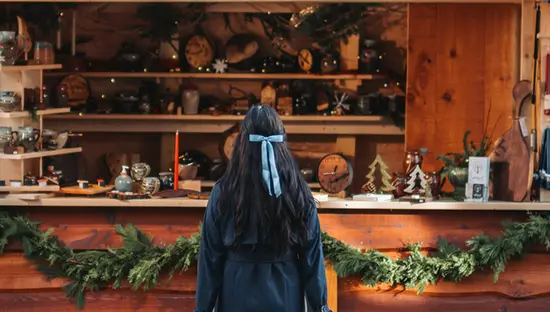 Jeune fille aux cheveux noirs foncés avec boucle bleue regardant vers un stand de marché des Fêtes où sont exposées des décorations en bois.
