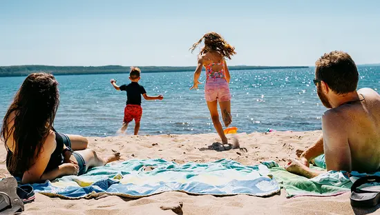 Two adults relax on towels at a sandy lakeside beach while two children run toward the water on a sunny day with clear blue sky.  
