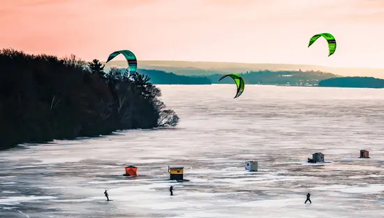Menschen beim Eiskiten und Eisfischen auf dem zugefrorenen Rice Lake unter einem rosafarbenen Abendhimmel.