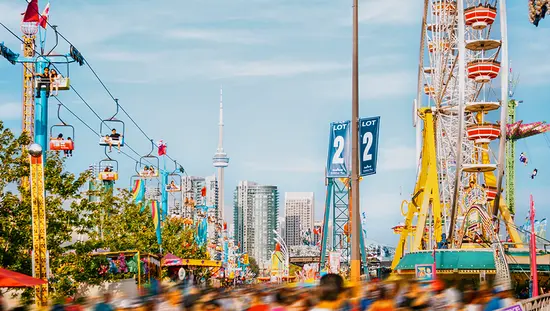 Crowds walk beside a sky ride, midway games, and the Toronto skyline, including the CN Tower, in the background.