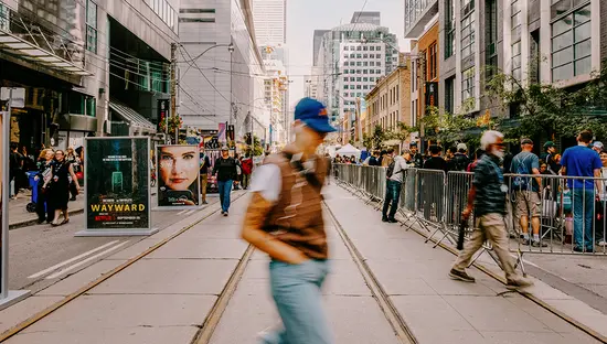 Pedestrians cross a downtown streetcar corridor lined with tall buildings, crowds and advertising boards.