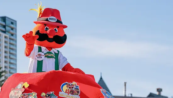 Oktoberfest mascot in a red hat waves from a parade float in an urban setting.
