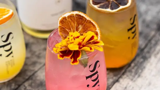 Three glasses of colourful cider topped with fruit and flowers sit on a wooden table.