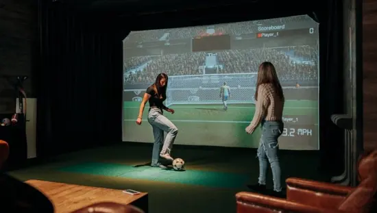 Un jugador tiene el pie sobre un balón de fútbol y está lanzando tiros a una portería virtual mientras otro jugador observa.