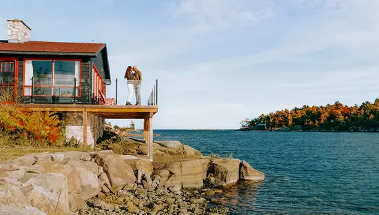 Lakeside cottage with a viewing deck overlooking calm water and a forested shoreline.
