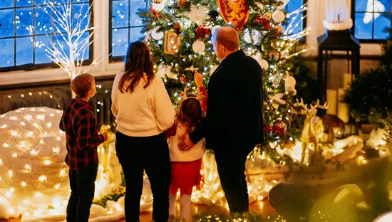 Eine Familie bewundert drinnen einen geschmückten Weihnachtsbaum, der von festlichen Lichtern und Ornamenten umgeben ist.
