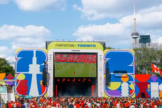 Foule rassemblée autour d’un grand écran extérieur diffusant un match de soccer lors d’un festival, avec la tour CN visible à l’arrière-plan.