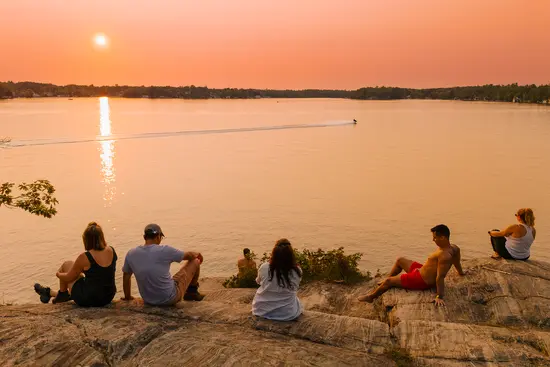 Des gens se détendant sur une rive rocheuse au bord d’un lac au coucher du soleil, avec une embarcation sur l’eau.