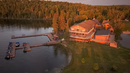 A lakeside lodge surrounded by autumn trees, with docks extending into the calm water and several boats moored along the shoreline.