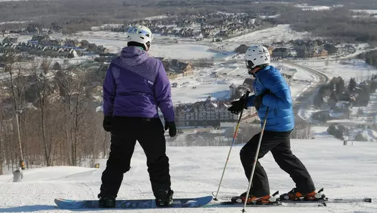 Two people at the top of a snowy hill looking down onto Blue Mountain Resort. One person has a snowboard and the other has skis.