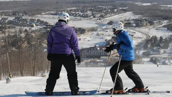 눈 덮인 언덕 꼭대기에 있는 두 사람이 블루 마운틴 리조트를 내려다보고 있습니다. 한 사람은 스노보드를 가지고 있고 다른 한 사람은 스키를 가지고 있습니다.