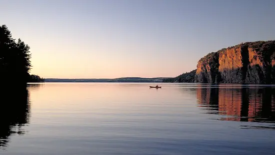 Cliffs overlook a serene lake at dusk with one lone kayaker.