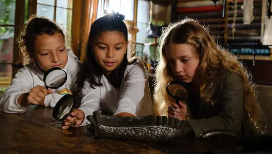 3 人の少女が拡大鏡を使って古い品物を調べています。