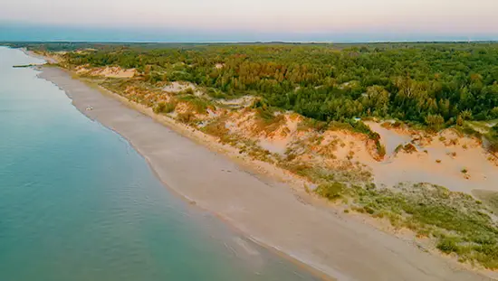 Aerial view of Ipperwash Beach in Ontario at dusk, with soft waves lapping a long stretch of shoreline backed by sandy dunes and dense forest.