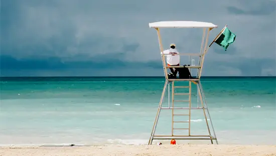 Lifeguard sitting on a tall white chair overlooking the turquoise waters of Grand Bend Beach, with a green flag fluttering in the breeze.