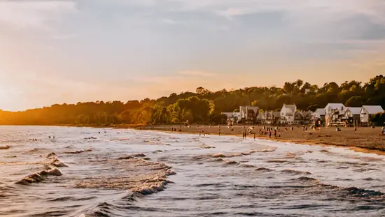 Waves rolling toward Port Stanley Beach, where swimmers and sunbathers enjoy the sunset, with charming cottages and tree-lined surroundings.