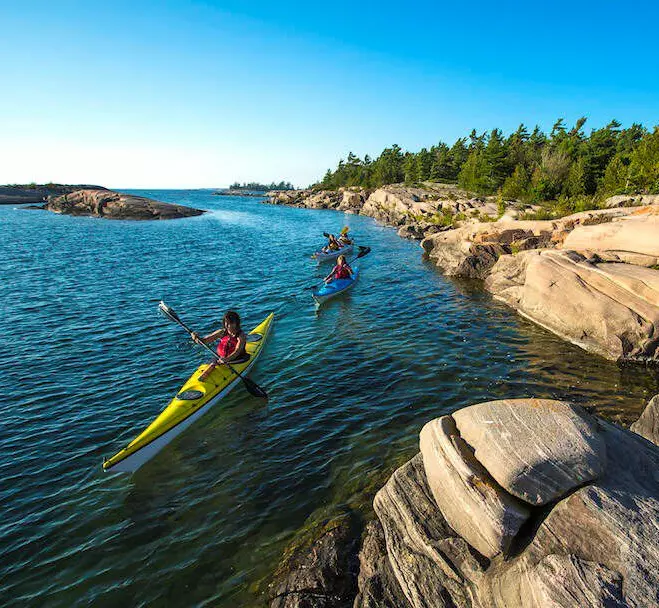 세 사람이 나무가 있는 바위 땅 근처의 호수를 따라 노를 저어 가고 있습니다.