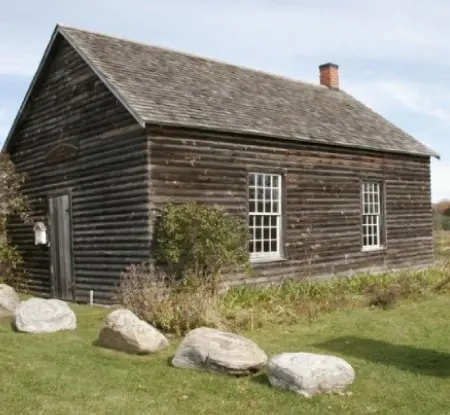Eine mit Steinen bedeckte Gedenktafel neben einem historischen Blockhaus.