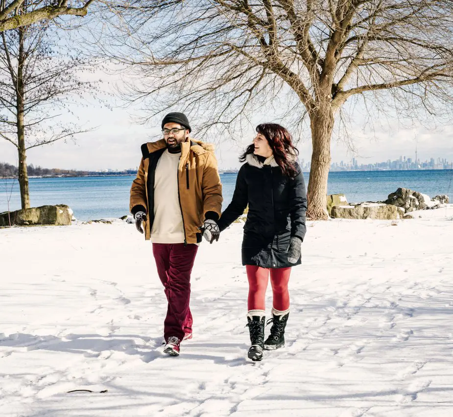 A man and a woman holding hands, walking on snow covered ground beside the lake.