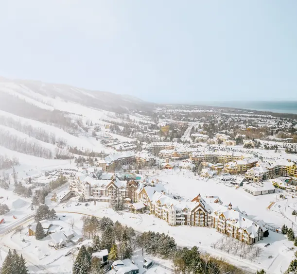 Aerial view of a snow-covered ski village with chalet-style buildings, tree-lined runs, and Georgian Bay visible in the distance.