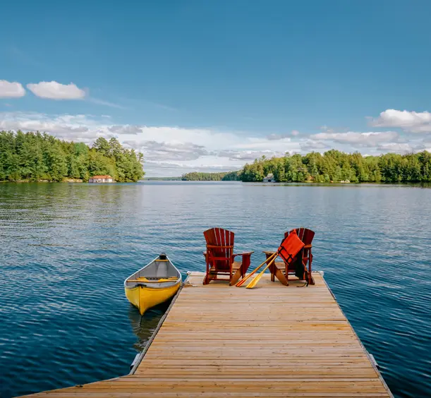 Ein Holzsteg mit Adirondack-Stühlen und einem gelben Kanu mit Blick auf einen ruhigen See und eine bewaldete Küste unter einem klaren blauen Himmel