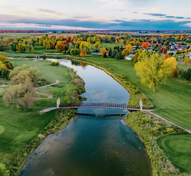 St. Jacobs et Elmira Scène automnale colorée d'une rivière serpentant à travers un terrain de golf, avec une passerelle piétonnière et une ville lointaine à l'horizon.