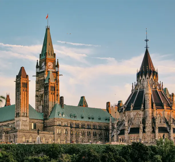 Looking out from Major’s Hill Park toward iconic gothic towers glowing in sunset light, framed by greenery and a soft blue sky.