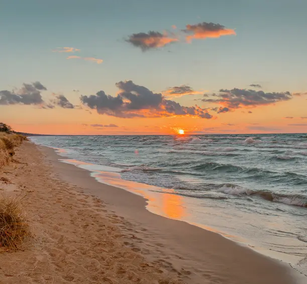 A sandy beach along Lake Huron at sunset, with the sun reflecting on the water and waves gently rolling ashore under a colourful sky.
