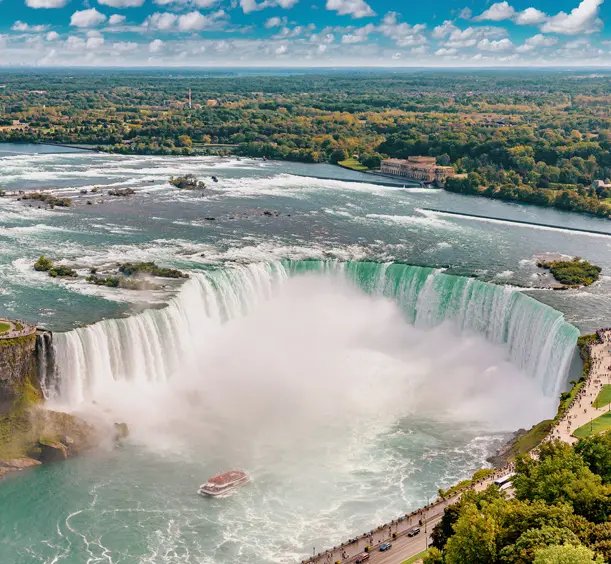 Aerial view of Horseshoe Falls on the Canadian side, with a sightseeing boat below and mist rising under a blue sky.