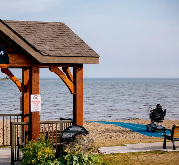 Holzpavillon mit Blick auf den Strand und das Wasser in Blind River, mit einem Elektromobil auf einem barrierefreien Weg, der zum Ufer führt.