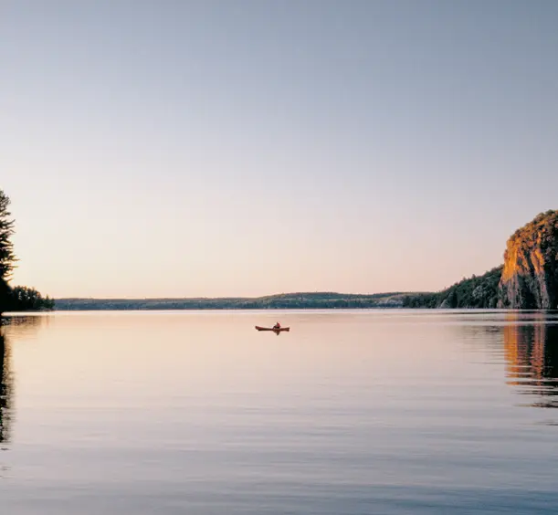 A person canoeing across a calm lake at sunset in Bon Echo Provincial Park, with forest on one side and a rocky cliff reflecting in the water.