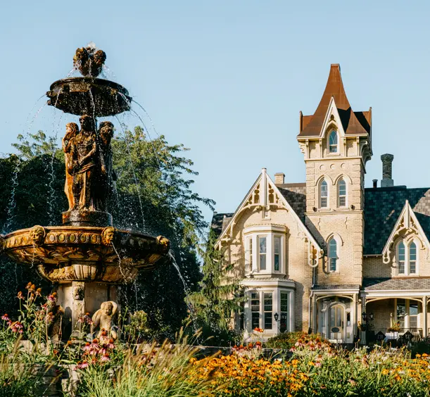 Victorian Gothic-style Elm Hurst Inn &amp; Spa, with a sculpted tiered fountain and vibrant garden blooms under a clear blue sky.