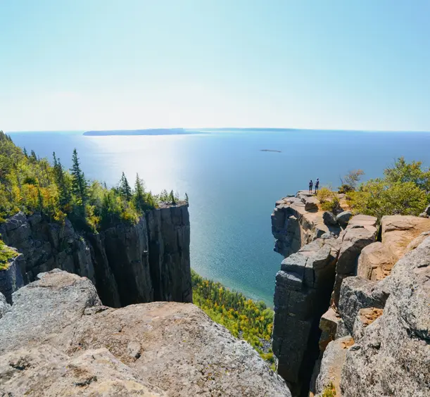 Two hikers standing on a rocky cliff overlooking Lake Superior at Sleeping Giant Provincial Park on the Sibley Peninsula, under a bright blue sky