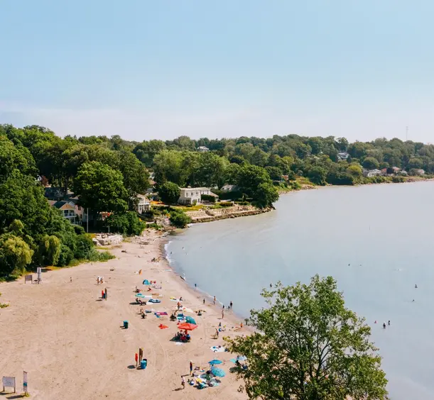 Beachgoers relaxing along the sandy shoreline of Lake Erie in Port Stanley, with calm waves and cottages nestled among lush trees.
