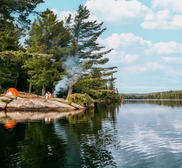 A lakeside campsite on a rocky shoreline in Algonquin Park, with an tent, a small campfire spewing smoke, and people gathered among tall pine trees.