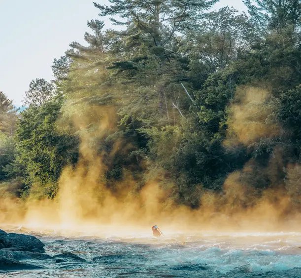Paddler navigating misty rapids on the Madawaska River in the Haliburton Highlands, surrounded by dense forest and early morning light.