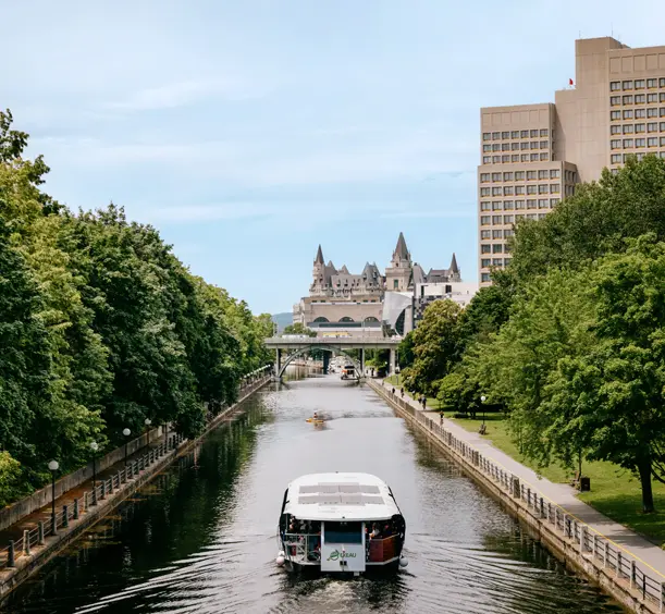 A boat cruising down the tree-lined Rideau Canal in Ottawa, with Parliament and a high-rise visible in the distance.